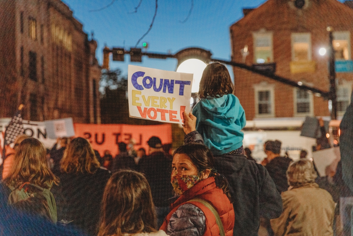 a feminine person holding a ‘count every vote’ sign among a group of people at a ‘count every vote’ rally at Penn Square.