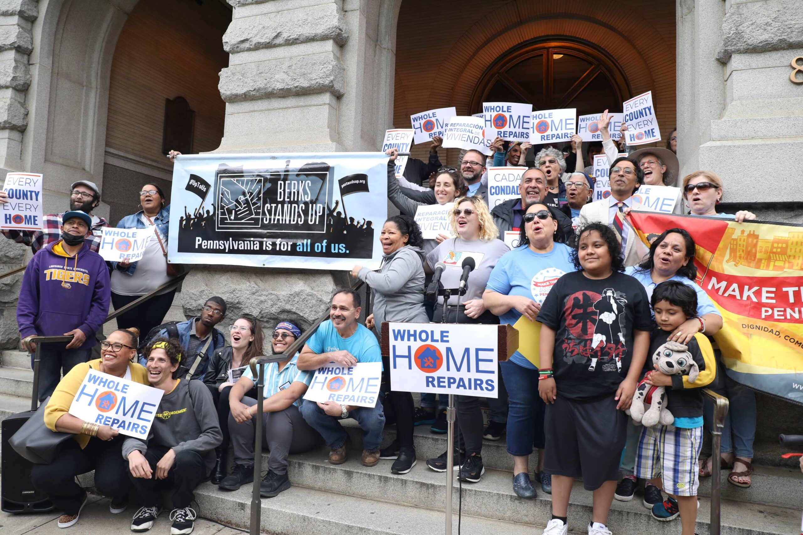 Group of people at the steps of a building smiling and holding signs that read "Whole Home Repairs".